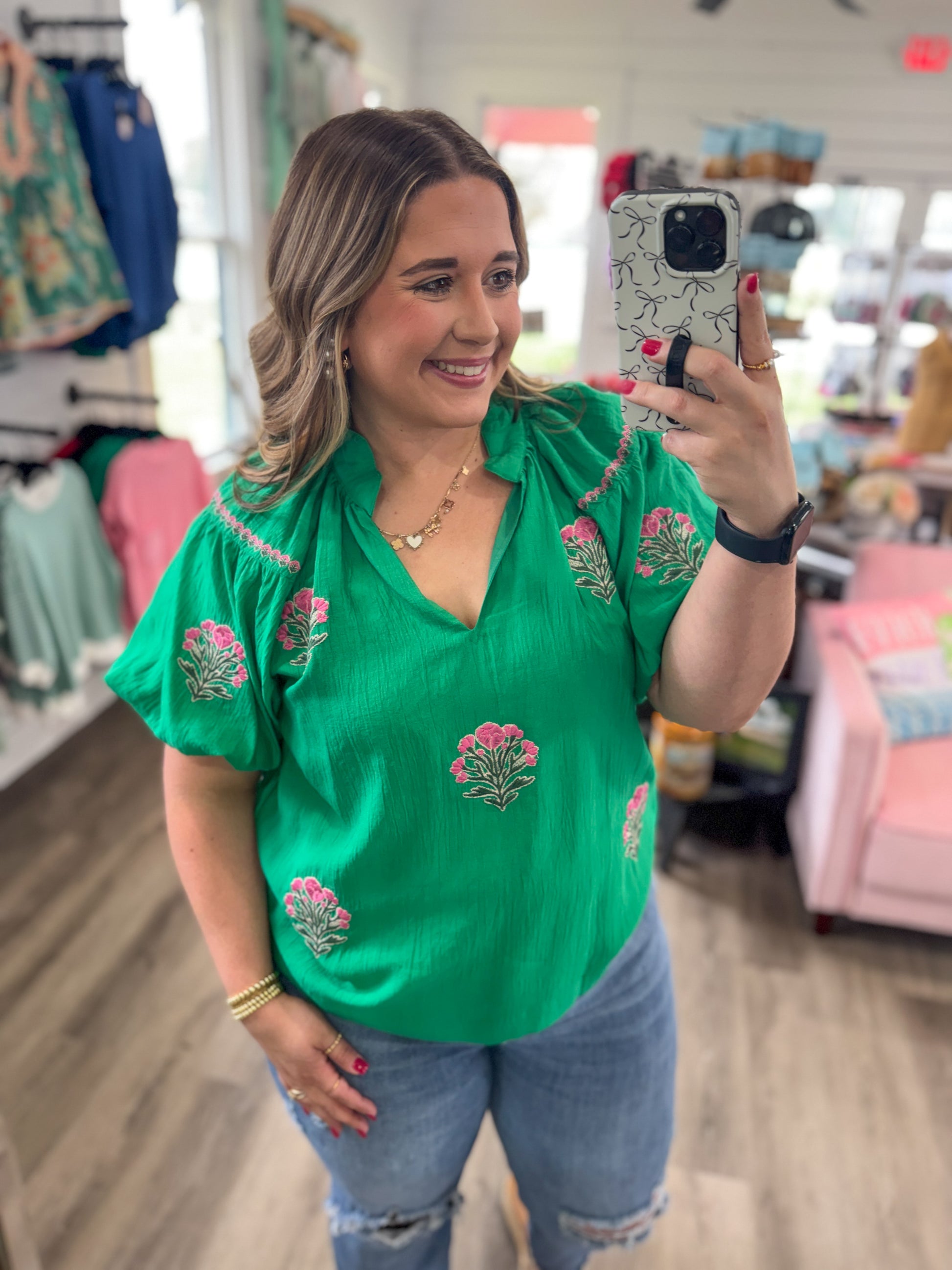 Woman taking a mirror selfie wearing a green embroidered top in a store.