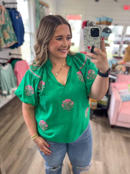 Woman taking a mirror selfie wearing a green embroidered top in a store.
