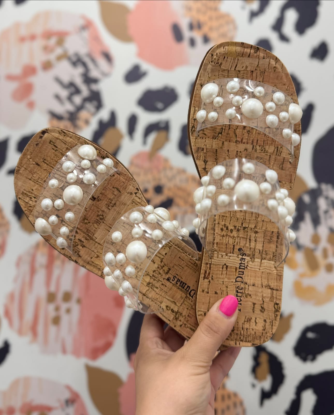A pair of cork-look flat sandals with clear straps decorated with white faux pearls, held by a hand with pink nail polish against a patterned wallpaper background.