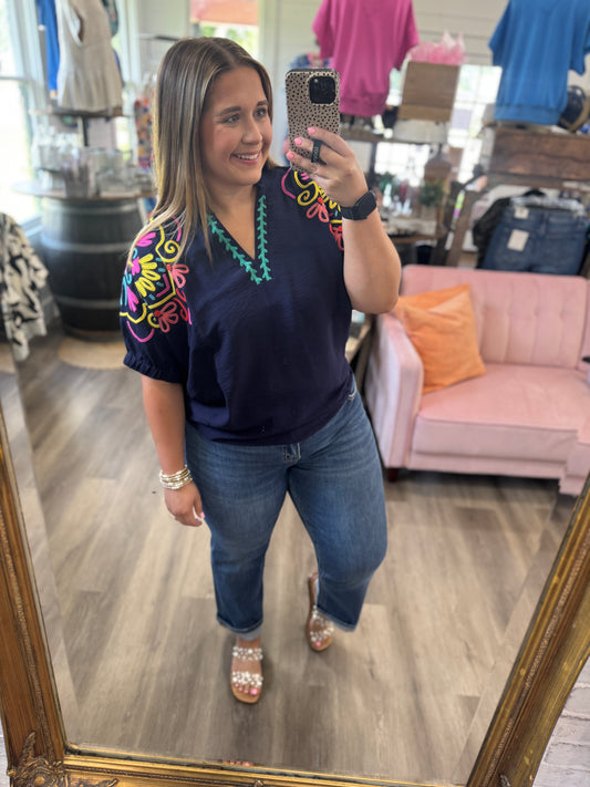 A woman takes a mirror selfie in a clothing boutique wearing a navy blue short-sleeve top with colorful embroidered details on the shoulders and a turquoise V-neck trim, paired with blue jeans.