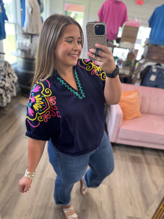 A woman takes a mirror selfie in a boutique wearing a navy blue short-sleeve blouse with colorful embroidered details on the shoulders and neckline, paired with blue jeans.