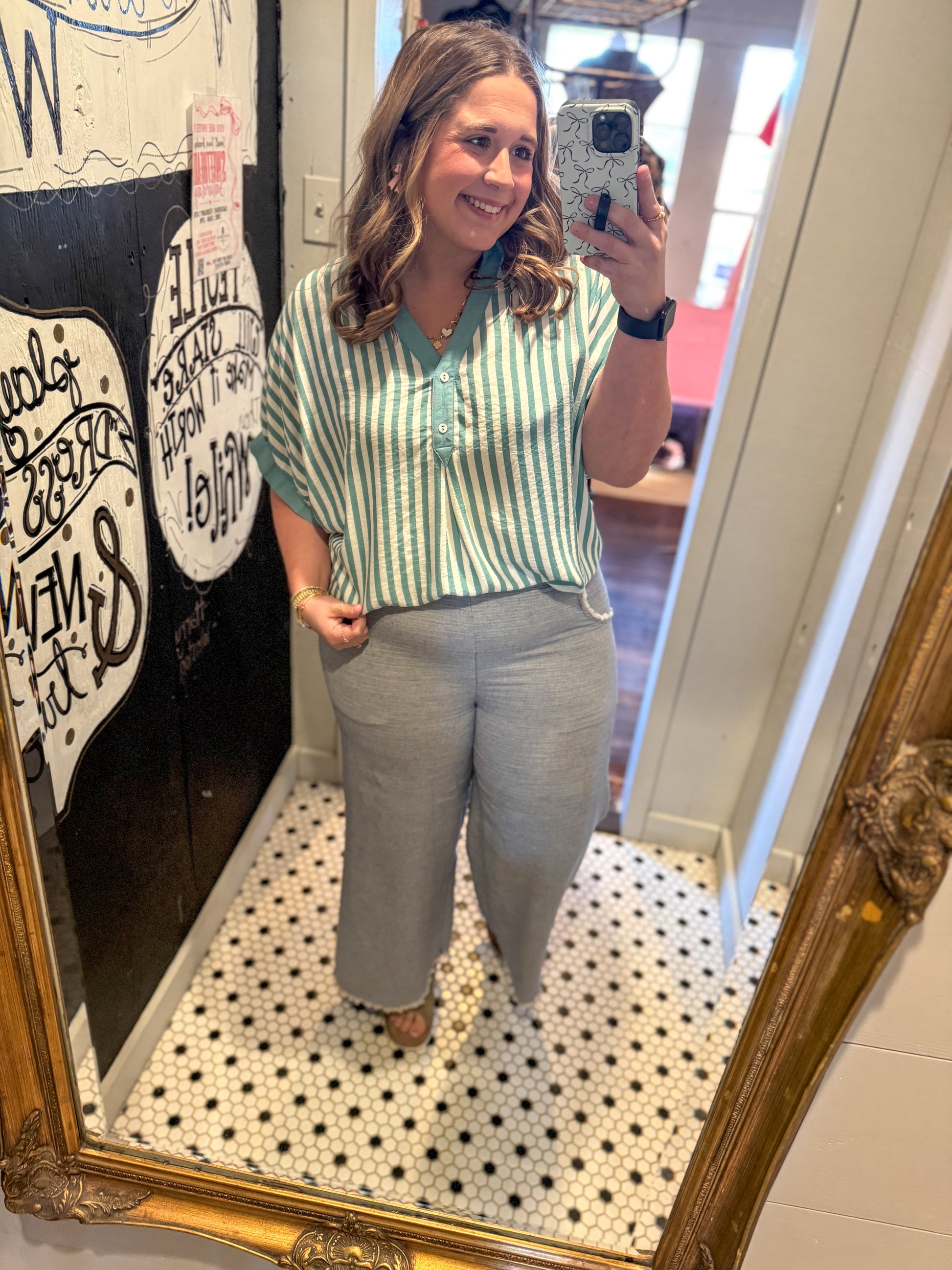 Woman taking a mirror selfie wearing a green and white striped shirt and gray pants.