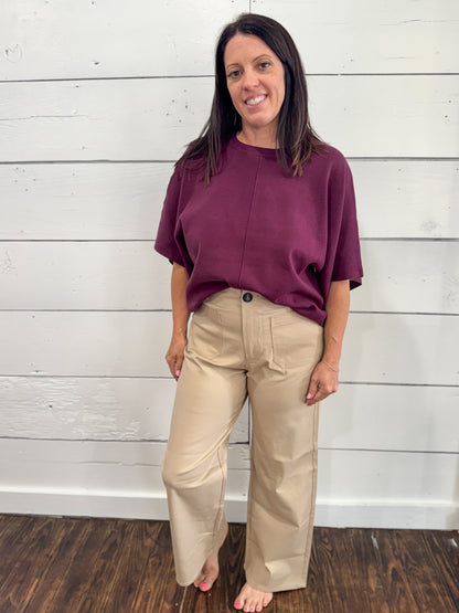 A woman stands barefoot against a white shiplap wall, wearing a loose short-sleeve burgundy top and beige wide-leg pants.