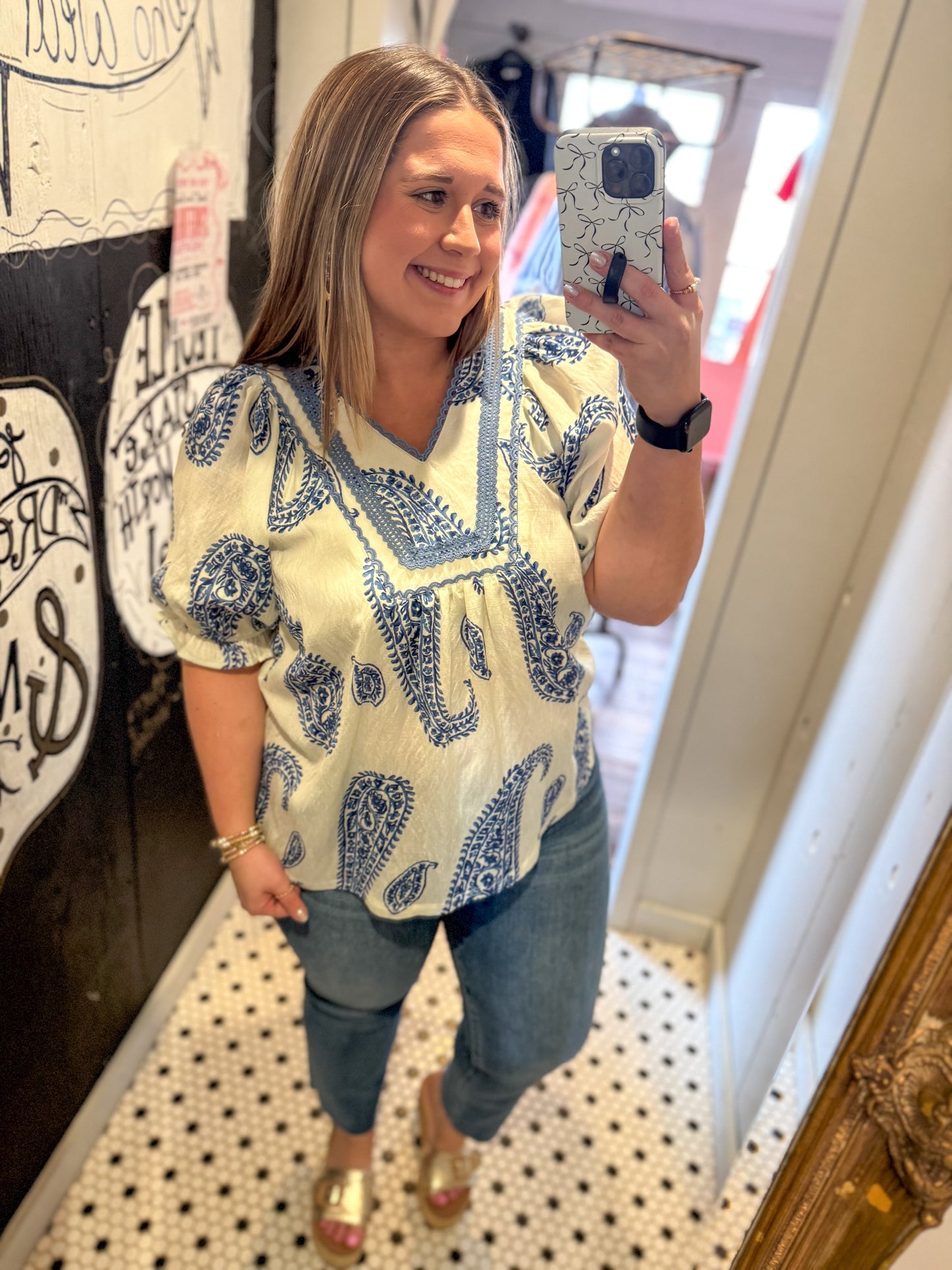 Woman taking a mirror selfie wearing a patterned top and jeans indoors.