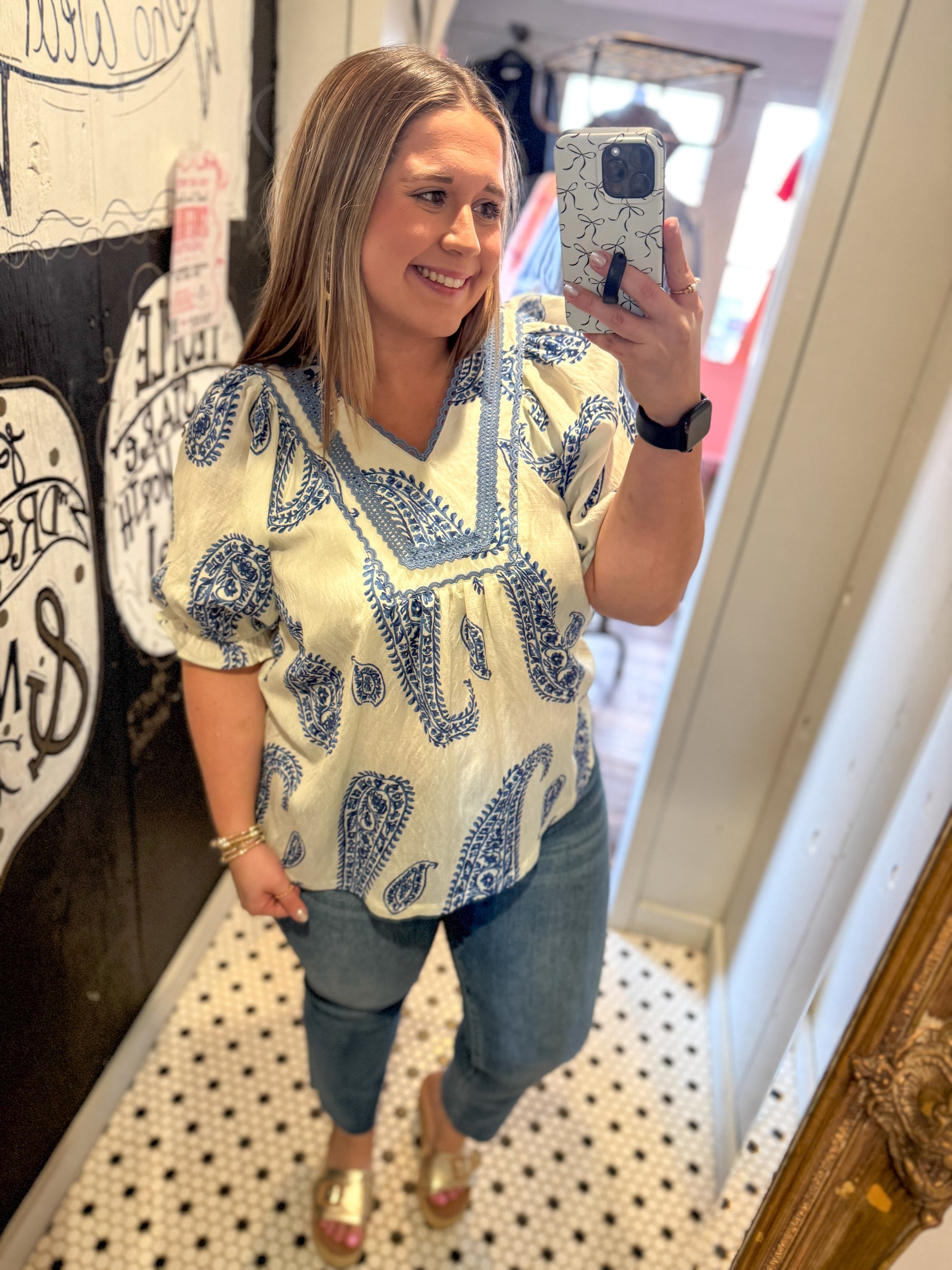 Woman taking a mirror selfie wearing a patterned top and jeans indoors.
