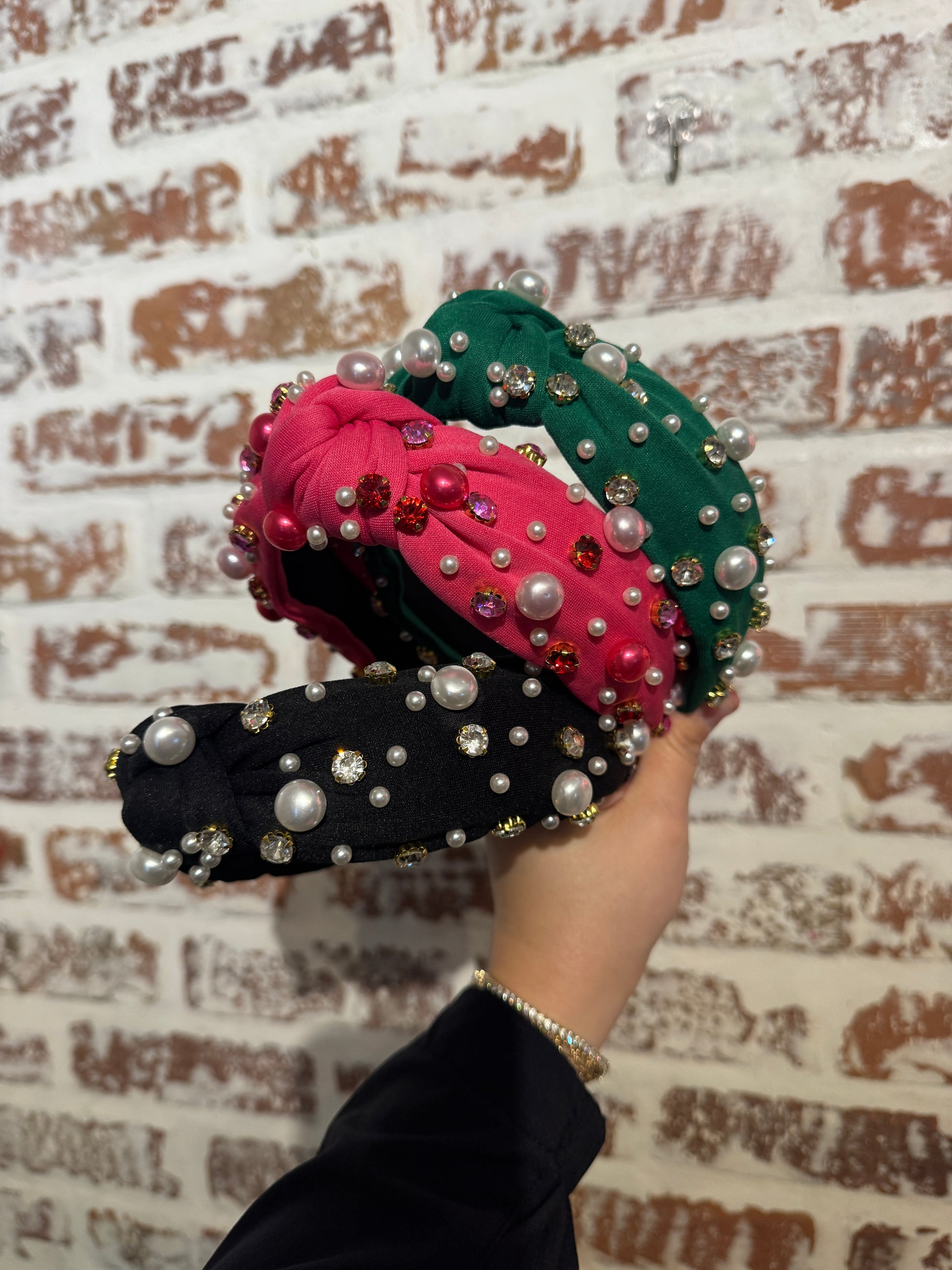 Three decorative headbands in green, pink, and black held by a hand against a textured wall.