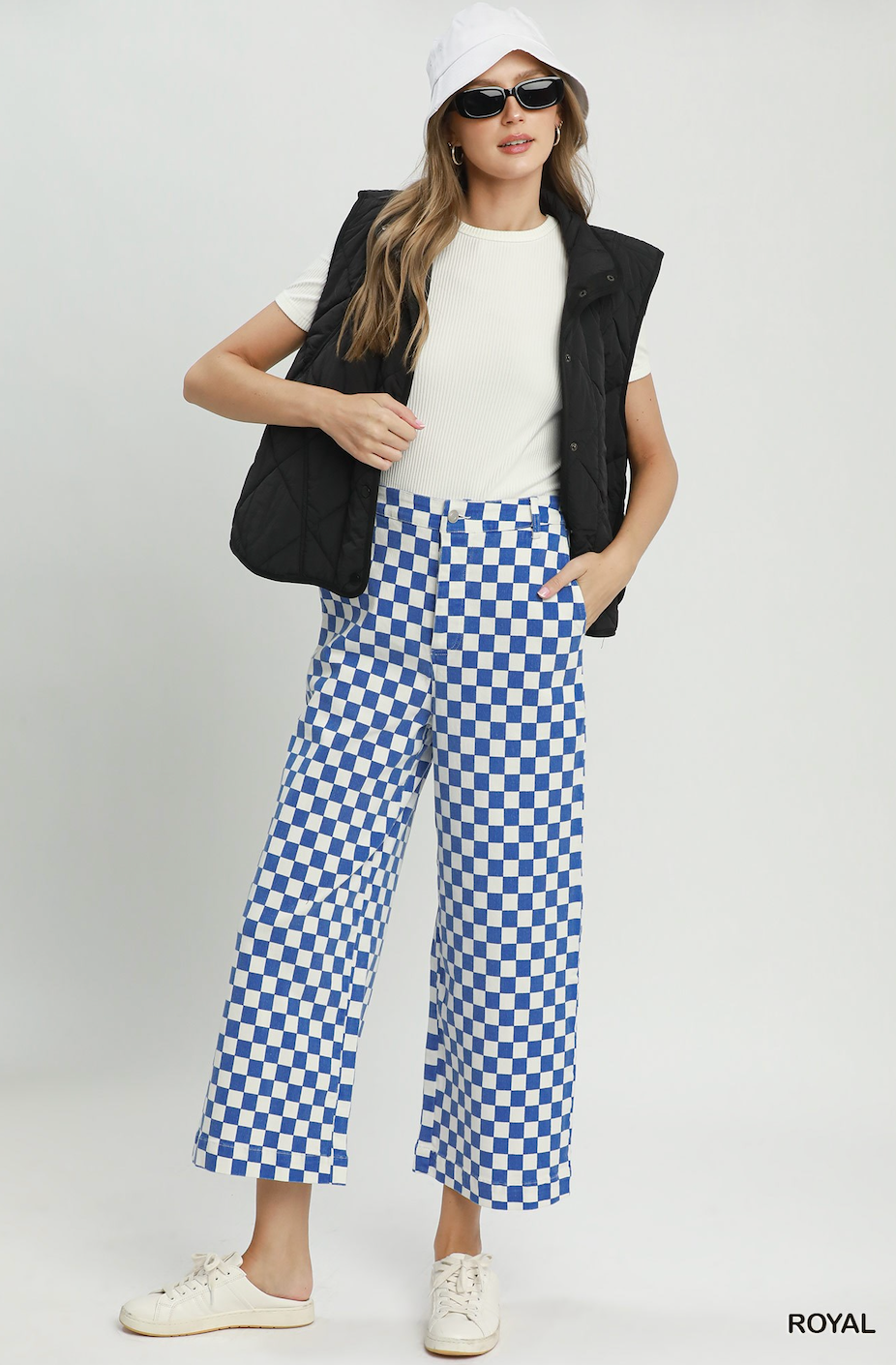 A model is shown wearing a stylish outfit featuring blue and white checkered wide-leg cropped pants, a white t-shirt, a black quilted vest, a white bucket hat, and white sneakers against a light gray studio background.