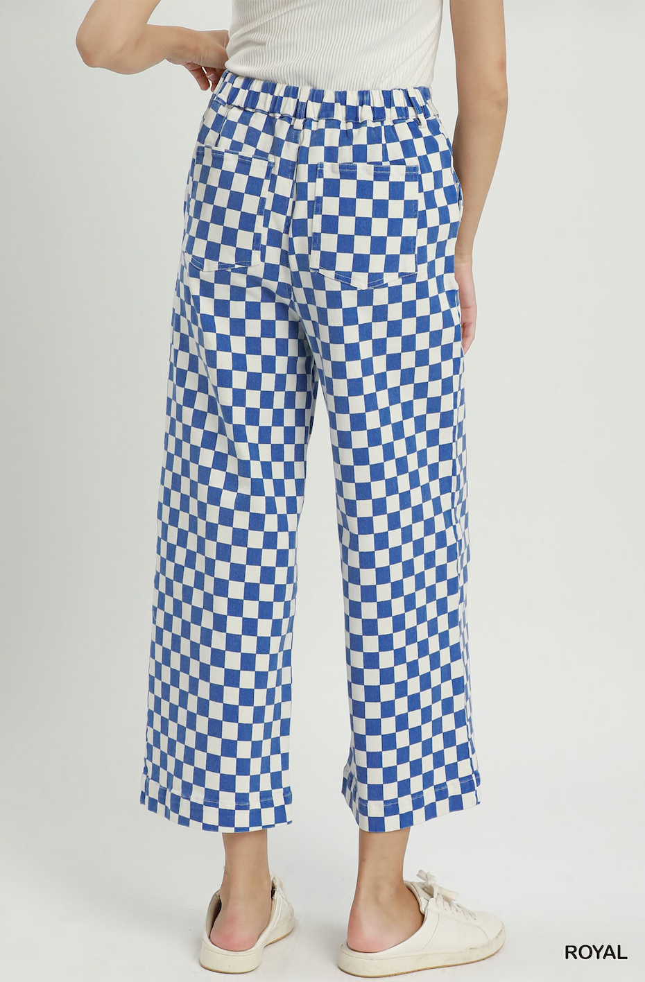 A person shown from the back wearing blue and white checkered cropped pants, a white ribbed top, and white slip-on shoes against a light gray studio background. The word ROYAL appears in the bottom right corner.