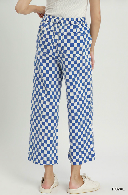 A person shown from the back wearing blue and white checkered cropped pants, a white ribbed top, and white slip-on shoes against a light gray studio background. The word ROYAL appears in the bottom right corner.