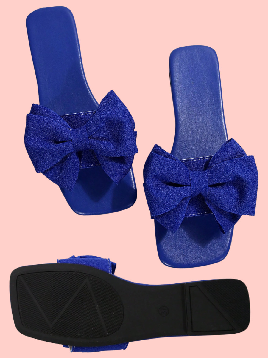 A pair of royal blue slide sandals with large bow details on the front, shown from the top and the sole, against a solid light pink background.