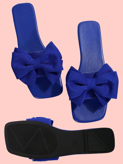 A pair of royal blue slide sandals with large bow details on the front, shown from the top and the sole, against a solid light pink background.