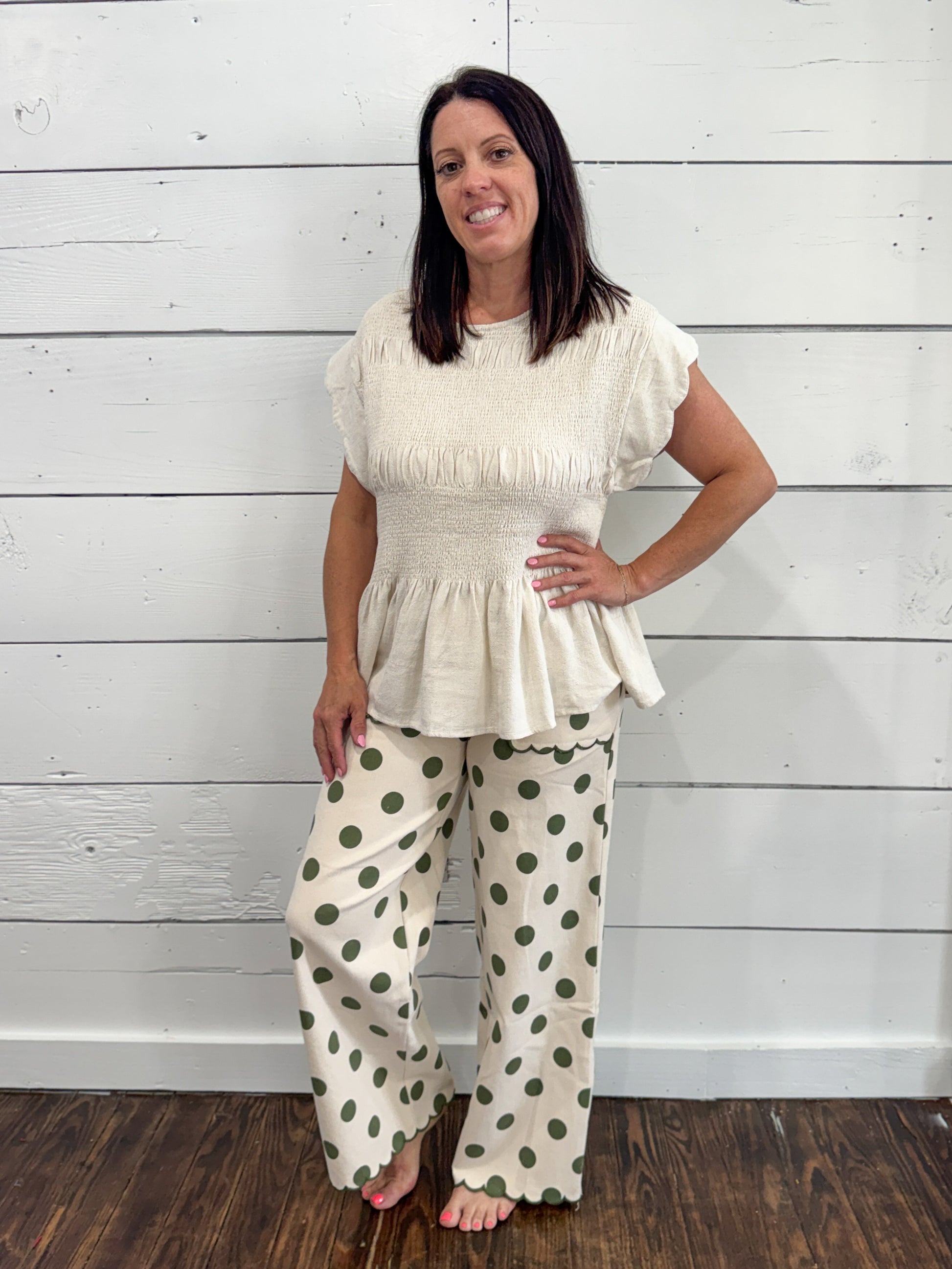 A woman modeling a cream-colored two-piece outfit consisting of a short-sleeve peplum top with fringe and matching wide-leg pants with green polka dots, standing against a white wood-paneled wall.