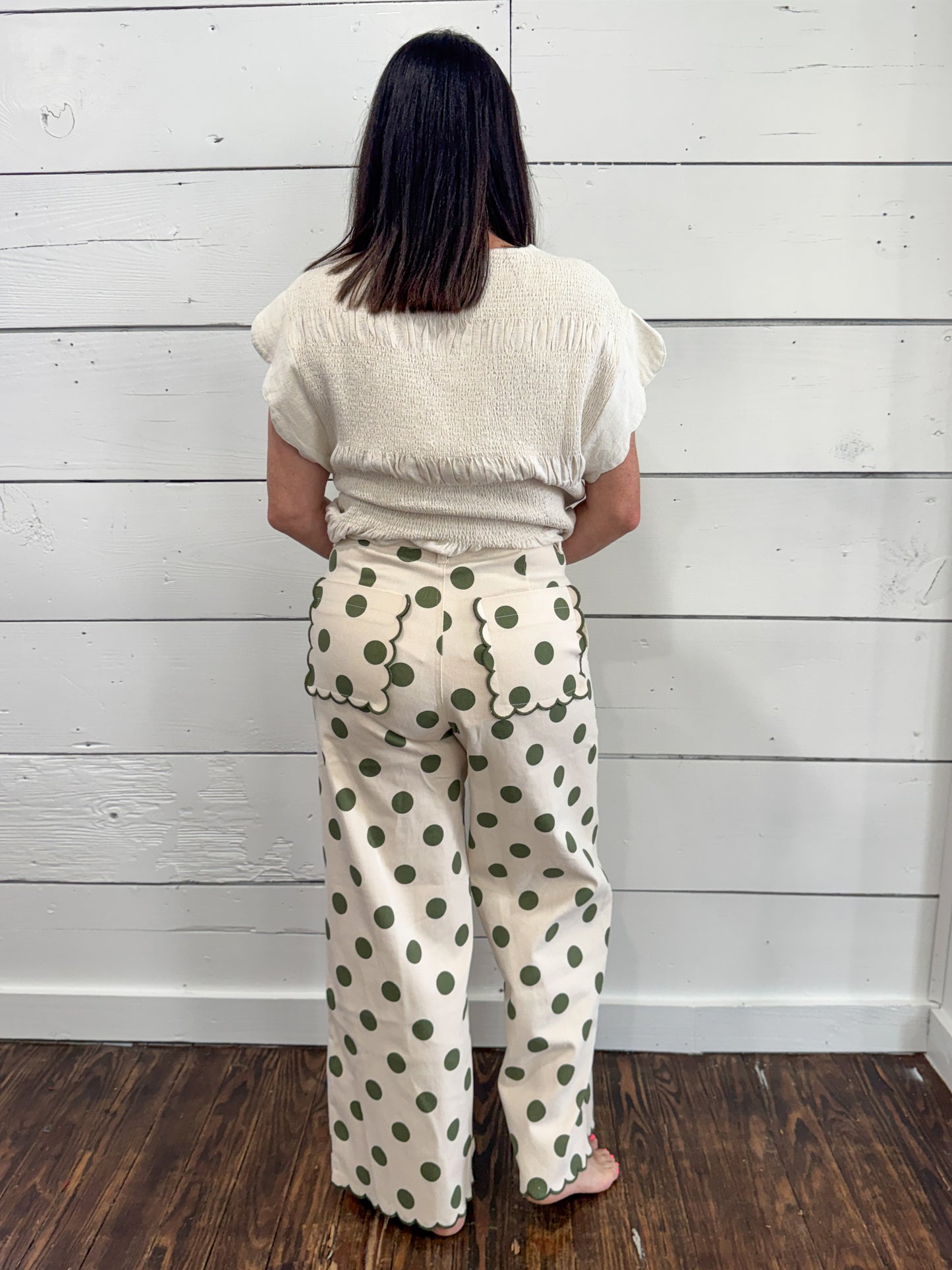 A woman viewed from behind wearing cream-colored wide-leg pants with a green polka dot pattern and a matching cream top, standing against a white wood-paneled wall on a dark wood floor.