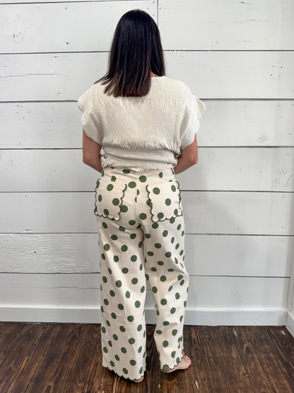 A woman viewed from behind wearing cream-colored wide-leg pants with a green polka dot pattern and a matching cream top, standing against a white wood-paneled wall on a dark wood floor.