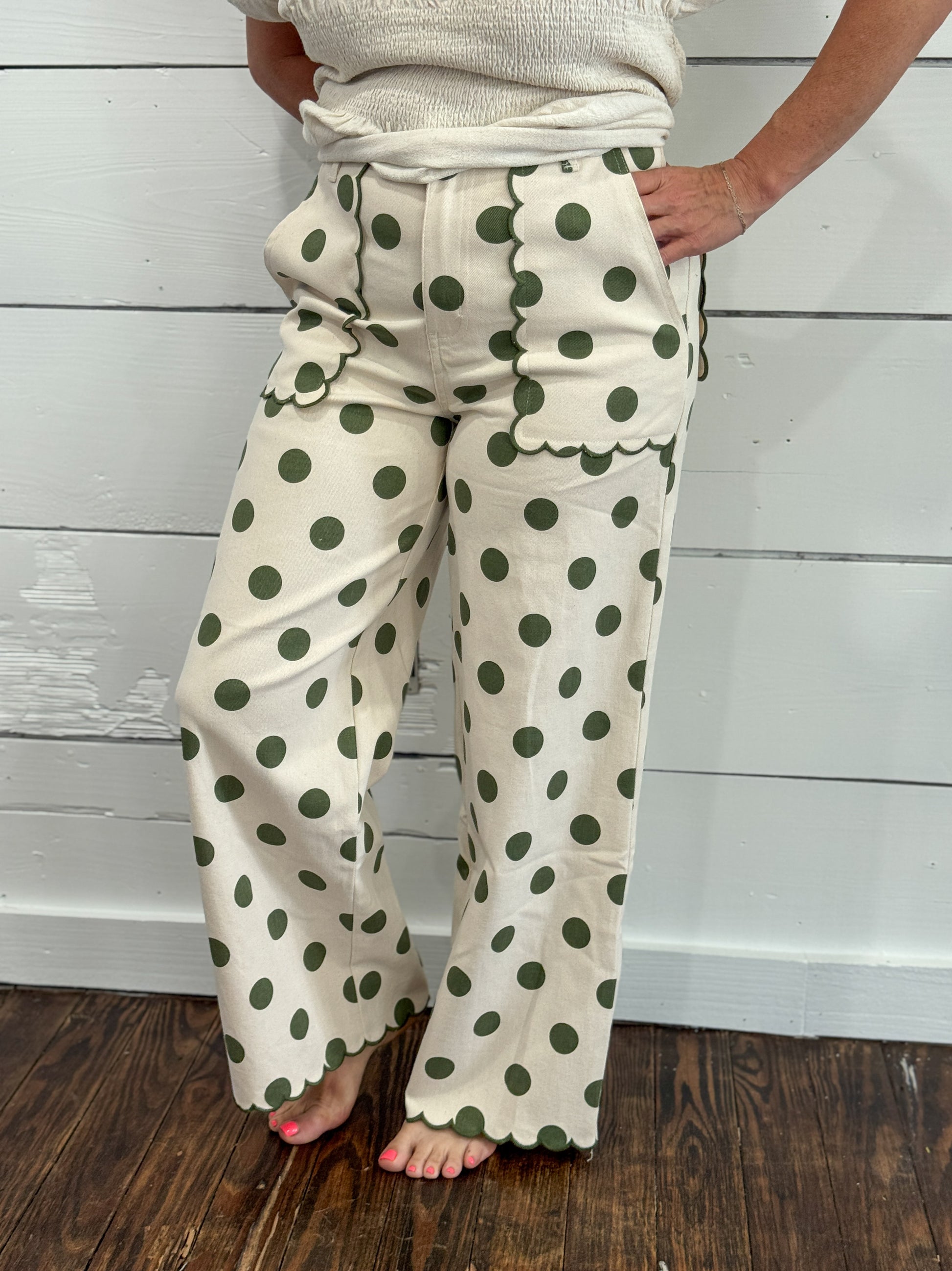 A person wearing cream wide-leg pants with dark green polka dots, standing barefoot on a dark wood floor against a white shiplap wall.