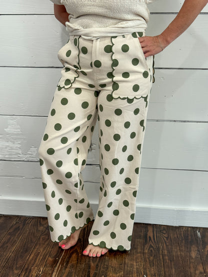 A person wearing cream wide-leg pants with dark green polka dots, standing barefoot on a dark wood floor against a white shiplap wall.
