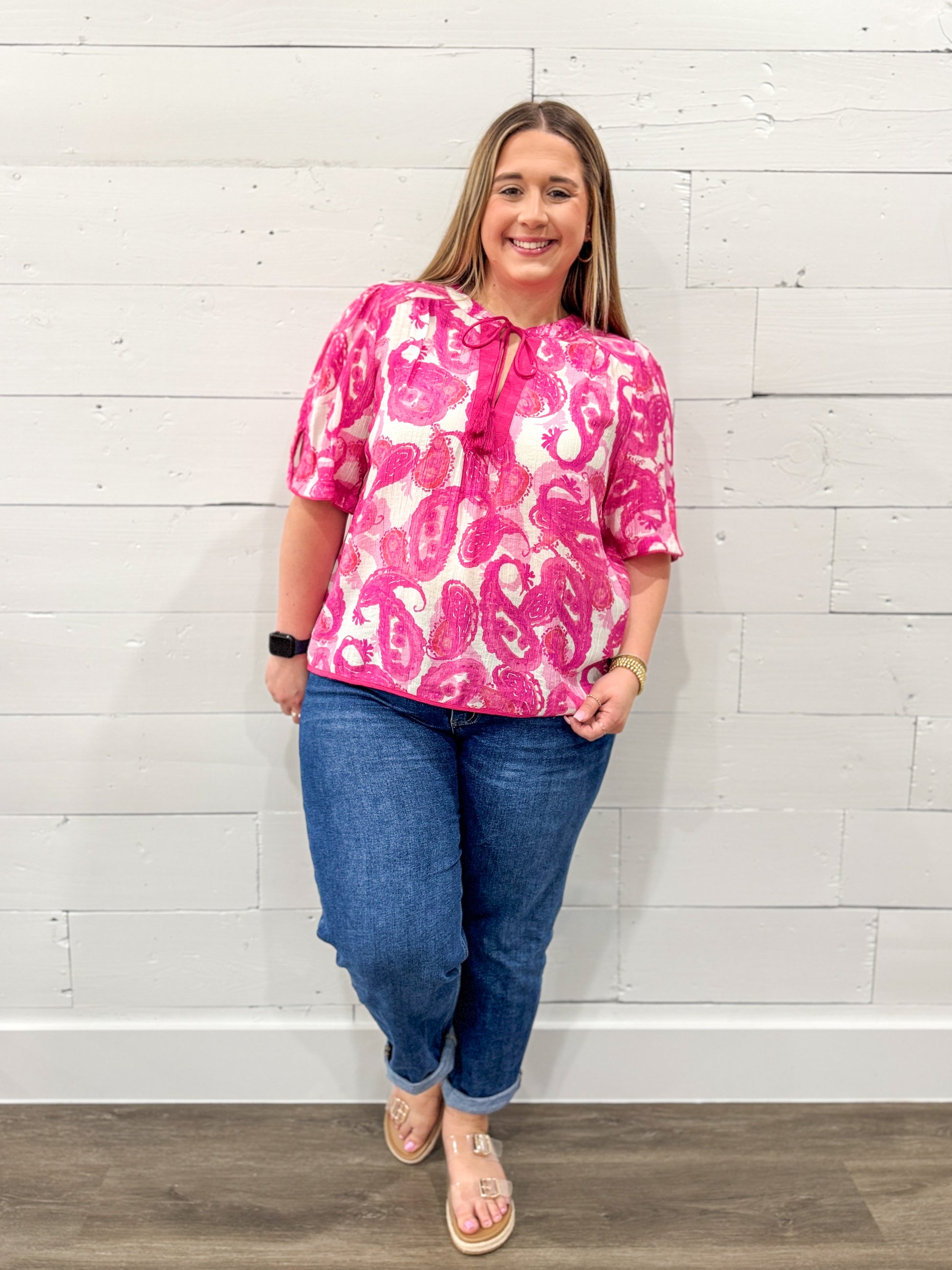 Woman wearing a pink patterned blouse and blue jeans against a white brick wall.