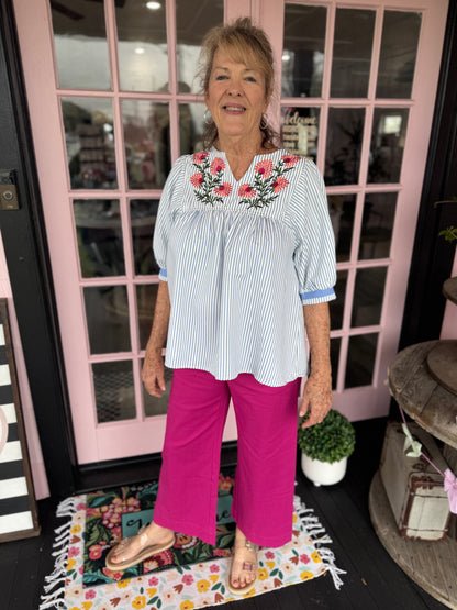 Woman wearing a white blouse with floral embroidery and pink pants standing in front of a glass door.