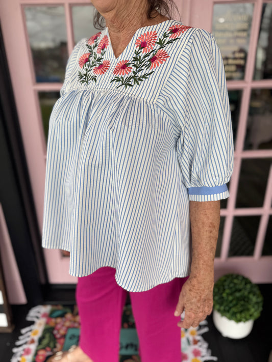 Person wearing a white blouse with floral embroidery and pink pants in front of a window.