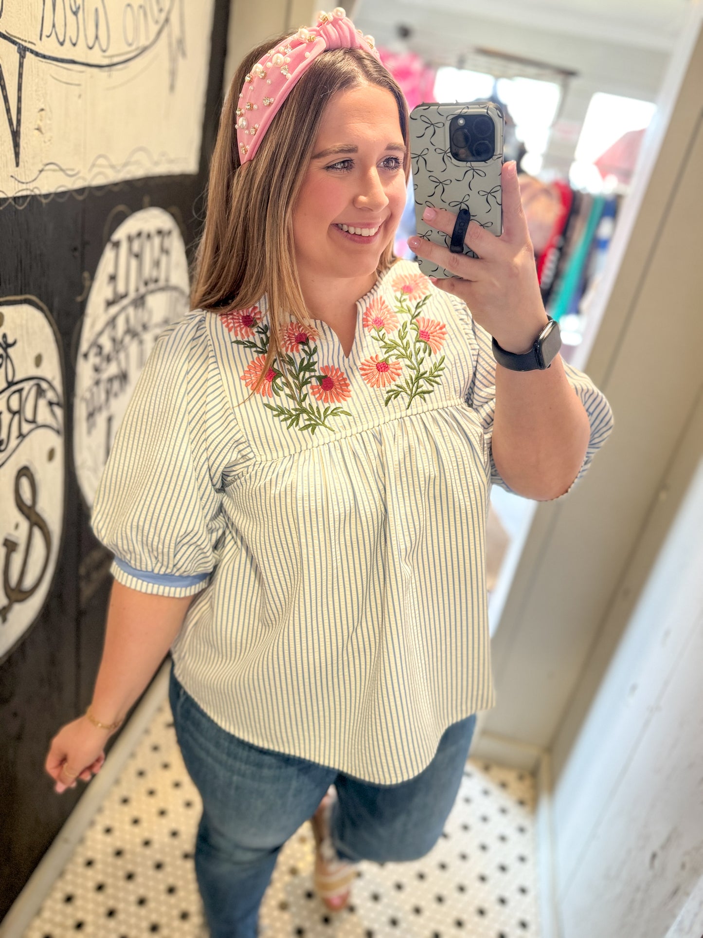 Woman taking a mirror selfie wearing a floral embroidered blouse and pink headband.
