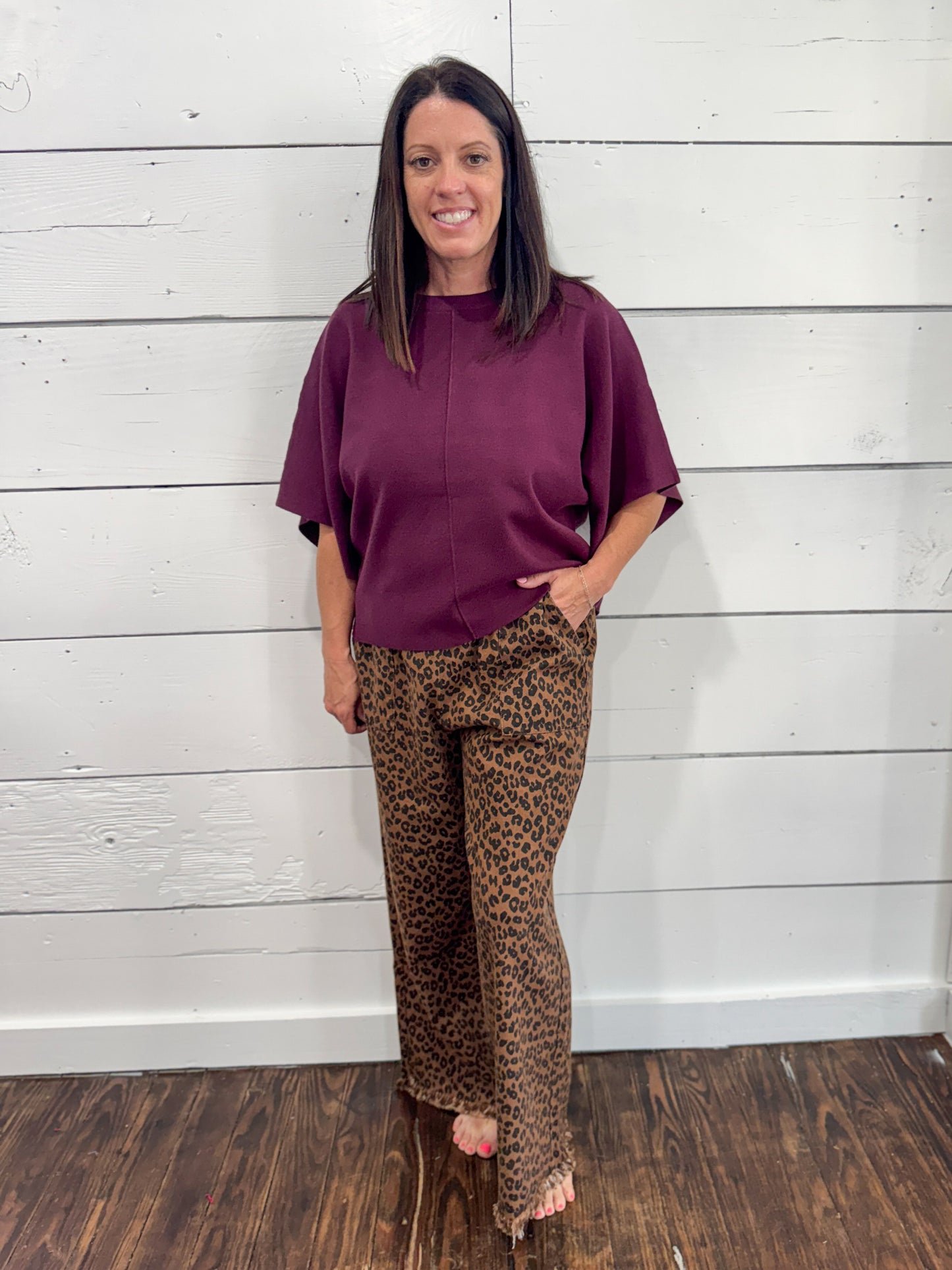 A woman modeling a loose-fitting short-sleeve top in deep plum purple paired with brown leopard-print pants, standing against a white wood-paneled wall.