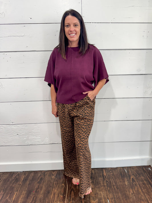 A woman modeling a loose-fitting short-sleeve top in deep plum purple paired with brown leopard-print pants, standing against a white wood-paneled wall.