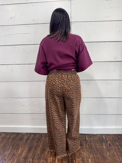 A person standing with their back to the camera, wearing a short-sleeve burgundy top and brown leopard-print pants, set against a white wood-paneled wall and dark wood floor.