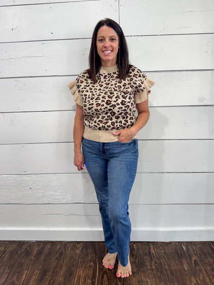 A woman modeling a beige and brown leopard print top with ruffled sleeves, paired with blue jeans, standing against a white shiplap wall.