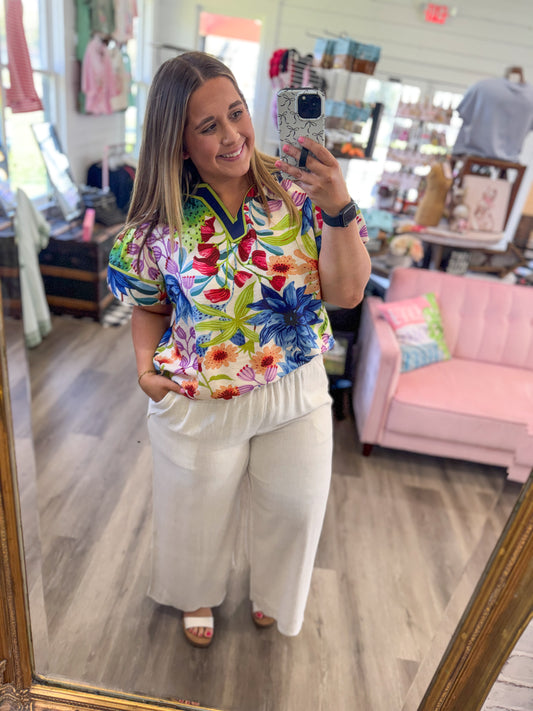 Woman taking a mirror selfie wearing a colorful floral shirt and white pants in a store.