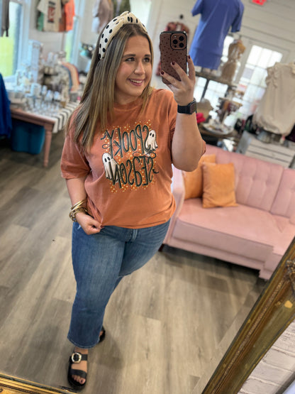 A woman takes a mirror selfie in a boutique wearing a peach-colored graphic t-shirt with 'Snoopy Halloween' written on it, blue jeans, and a black-and-white polka dot headband.