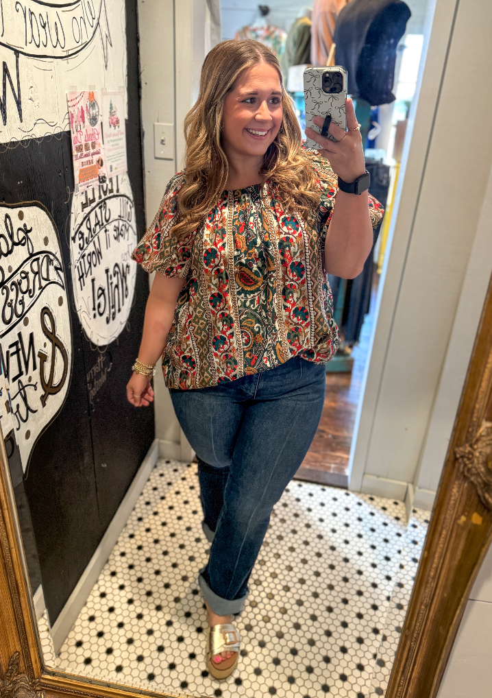 Woman taking a mirror selfie wearing a patterned top and jeans in a room with a decorative wall.