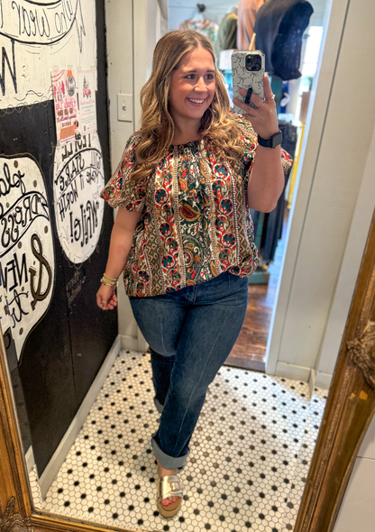Woman taking a mirror selfie wearing a patterned top and jeans in a room with a decorative wall.