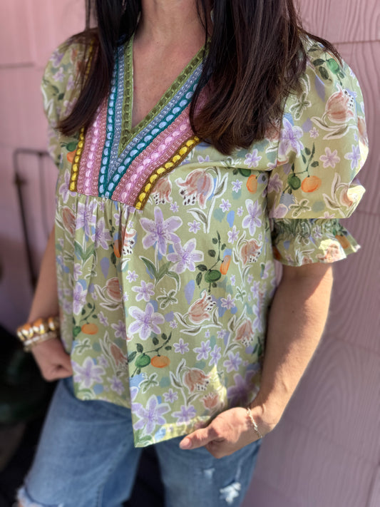 Person wearing a floral blouse with colorful embroidery against a pink wall.