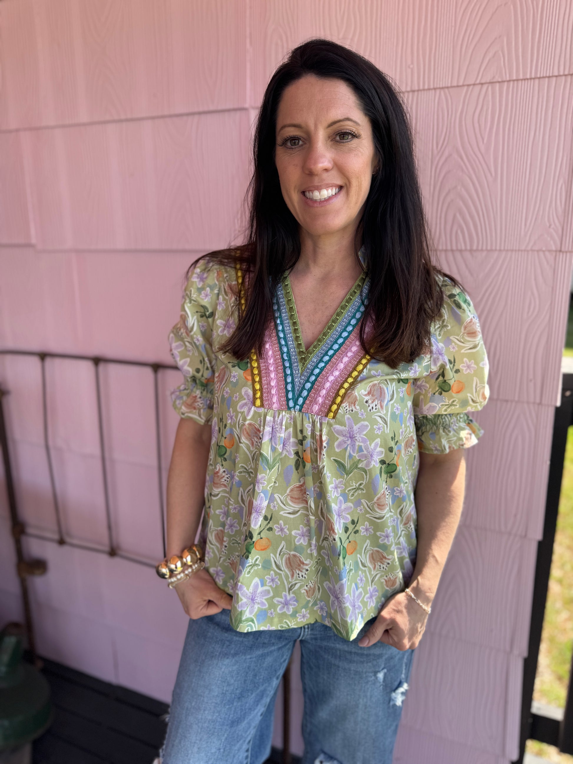 Woman wearing a floral blouse with colorful accents against a pink wall.