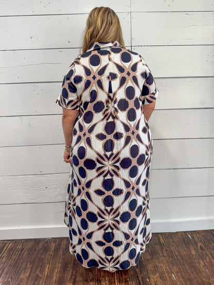 A woman viewed from behind wearing a short-sleeve collared dress with a geometric pattern in navy blue, beige, and white, standing against a white wood-paneled wall.