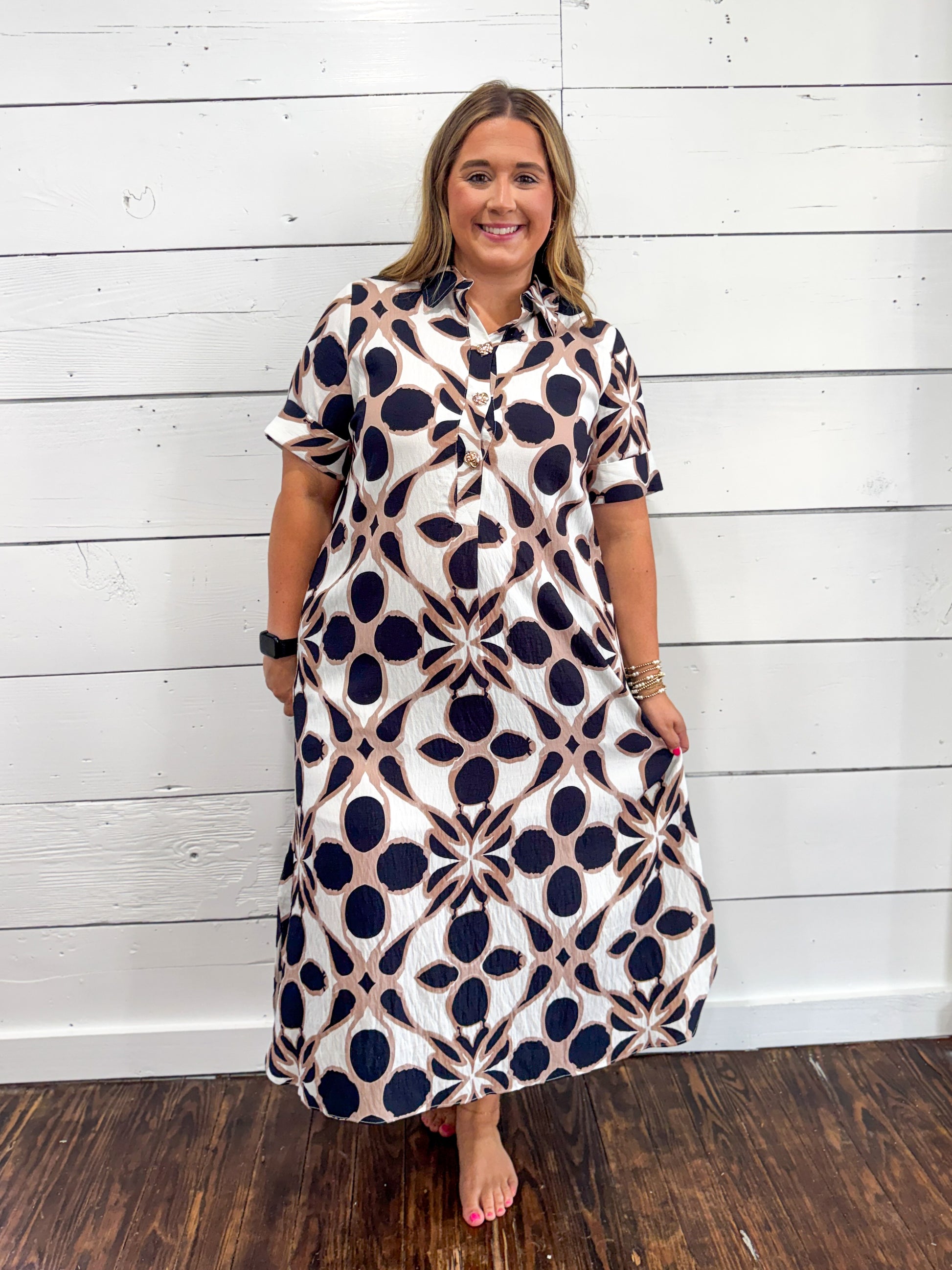 A woman modeling a short-sleeve button-front maxi dress with a geometric navy and beige pattern on a white background, standing against a white wood-paneled wall.