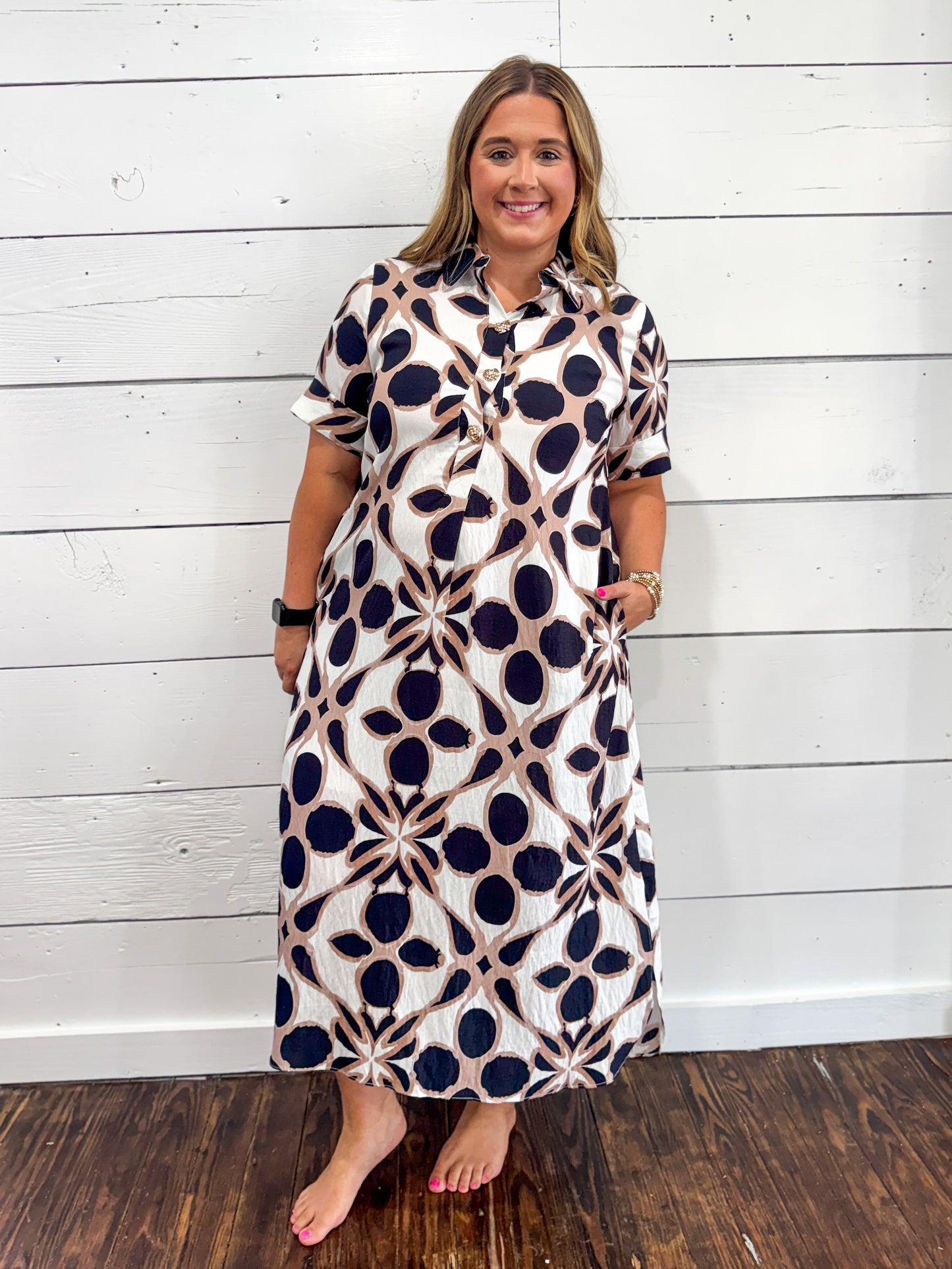A woman modeling a short-sleeve midi dress with a navy blue and beige geometric pattern on a white background, standing against a white wood-paneled wall.