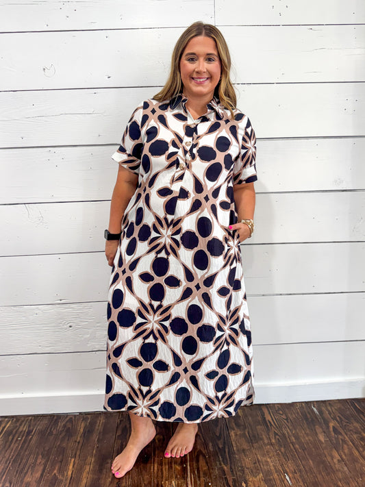 A woman modeling a short-sleeve midi dress with a navy blue and beige geometric pattern on a white background, standing against a white wood-paneled wall.