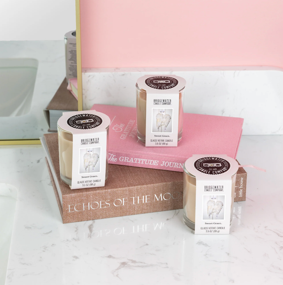 Three glass jar candles with white labels and gold wax tops are arranged on stacked books on a white marble surface, with a pink wall and mirror in the background.
