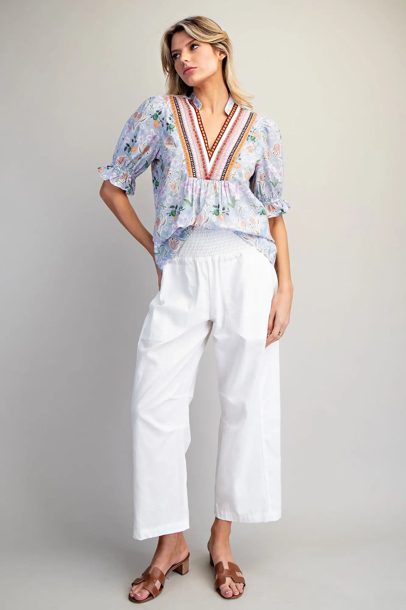 Woman wearing a floral blouse with colorful embroidery and white pants on a plain background