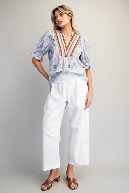 Woman wearing a floral blouse with colorful embroidery and white pants on a plain background