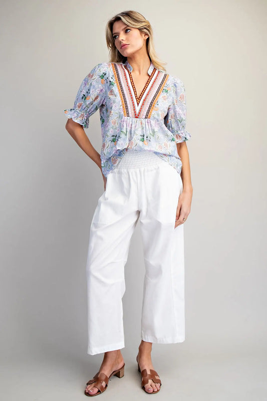 Woman wearing a floral blouse with colorful embroidery and white pants on a plain background
