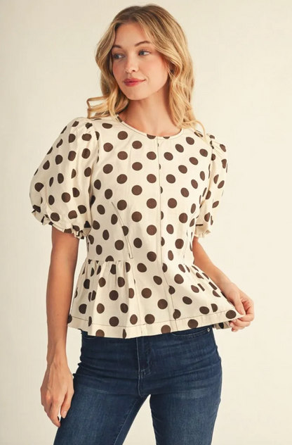 A woman wearing a cream-colored short-sleeve blouse with brown polka dots and puffed sleeves, paired with dark blue jeans, against a plain light beige background.