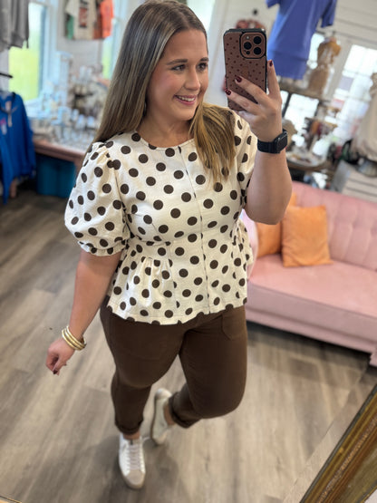 A woman takes a mirror selfie in a boutique while wearing a cream puff-sleeve blouse with brown polka dots and brown pants.