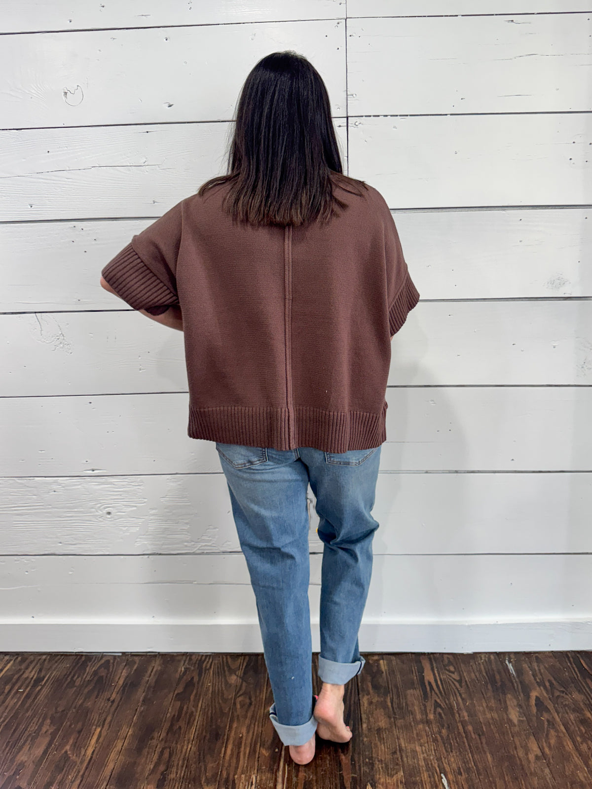 A person viewed from behind wearing a brown short-sleeve knit sweater and light blue jeans, standing barefoot on a dark wood floor against a white paneled wall.