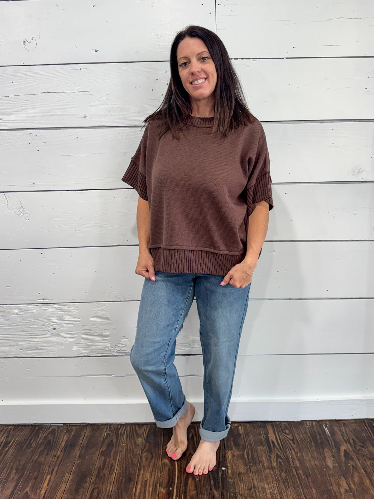 A woman modeling a brown short-sleeve knit top with ribbed trim, paired with blue jeans, standing against a white wood-paneled wall on a dark wood floor.