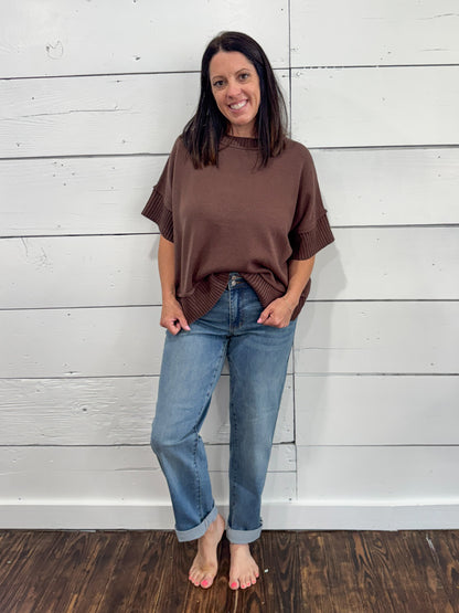A woman stands barefoot against a white wood plank wall, wearing an oversized brown short-sleeve top and light blue jeans.