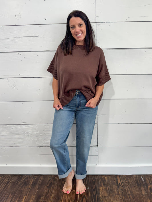 A woman stands barefoot against a white wood plank wall, wearing an oversized brown short-sleeve top and light blue jeans.