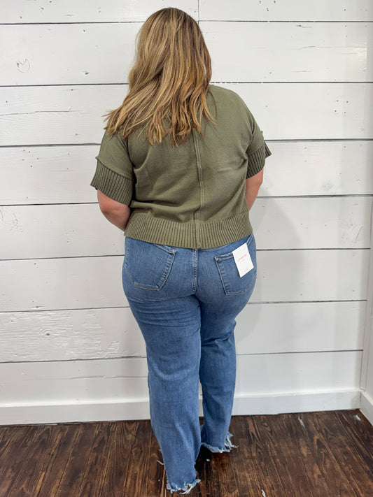 A woman viewed from behind wearing an olive green short-sleeve knit top and blue jeans, standing against a white wood-paneled wall on a dark wood floor.