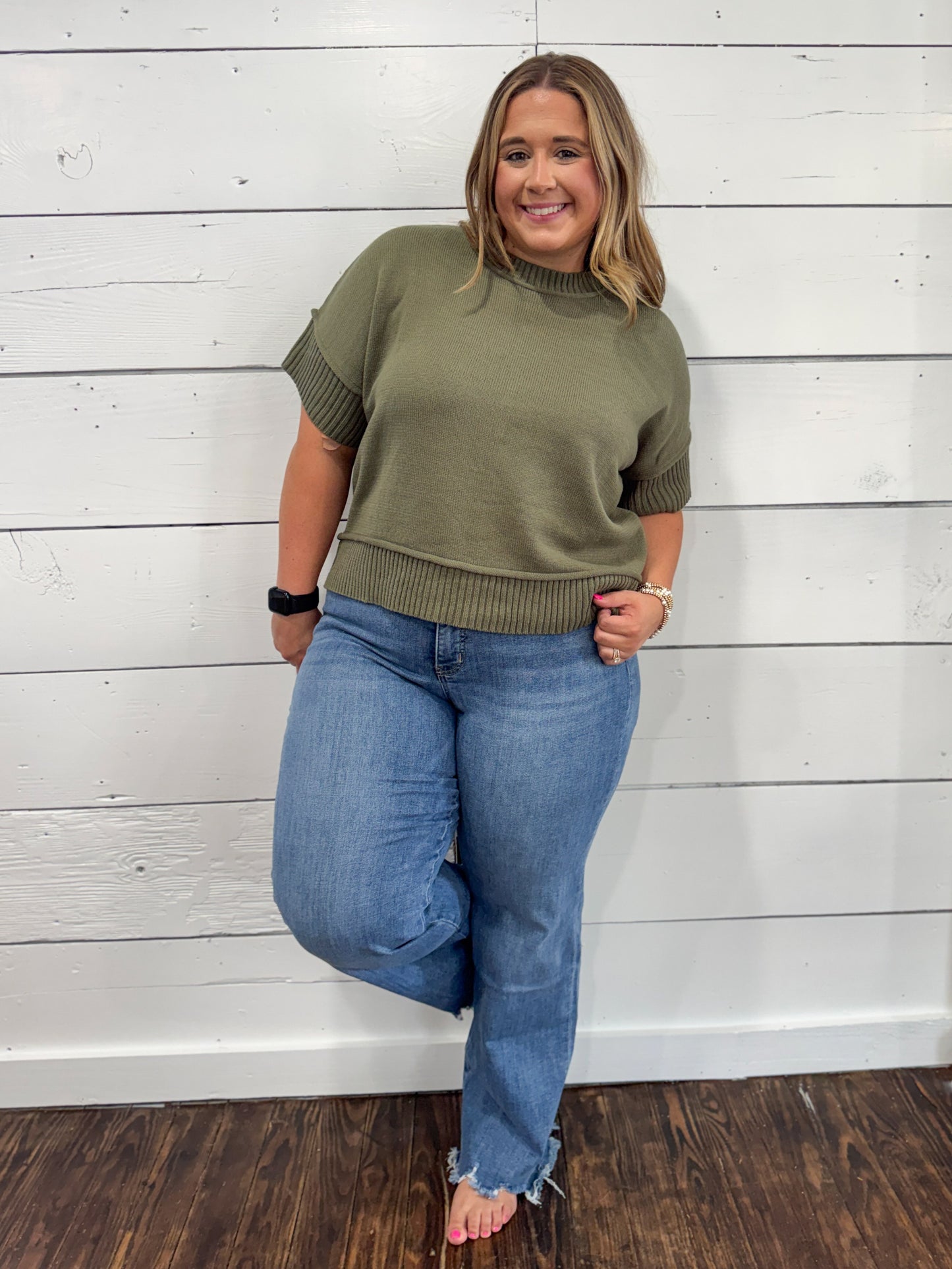 A woman modeling an olive green short-sleeve mock neck knit sweater with ribbed cuffs and hem, paired with blue jeans, standing against a white wood-paneled wall.